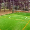 Campo sportivo con erba sintetica, righe arancioni e bianche, circondato da alberi.