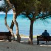 Uomo seduto su una panchina in spiaggia, alberi sullo sfondo e mare azzurro.