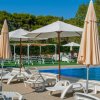 Piscina con lettini bianchi e ombrelloni su un patio, circondata da alberi verdi.