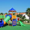 Area giochi colorata con scivoli e torrette su erba sintetica, cielo limpido e palme sullo sfondo.