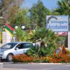 Ingresso di Villaggio Arcobaleno con cartello e auto davanti a un'aiuola fiorita.