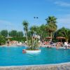 Piscina con centro vegetato, persone nuotano intorno. Palme e ombrelloni sul bordo.