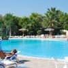 Piscina con donna che prende il sole su un lettino, ombrelloni e palme sullo sfondo.