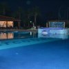 Piscina illuminata di notte con un edificio aperto e palme sullo sfondo.