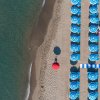 Vista aerea di una spiaggia con ombrelloni blu e un pedalò giallo sull'acqua.