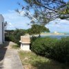 Vista su un vialetto costiero con edificio bianco, cespugli e mare azzurro in sottofondo.