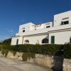 Edificio bianco moderno con finestre verdi, circondato da siepi e un muro di pietra.