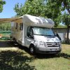 Camper bianco parcheggiato all'ombra di alberi, con sedie e tavolo sotto un telo esteso.