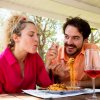 Due persone sorridenti sedute a un tavolo all'aperto, mangiano spaghetti e bevono vino.
