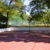 Campo da basket all'aperto ombreggiato da alberi, con un canestro e un campo da calcio sullo sfondo.