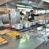 Cucina industriale con pentole in acciaio, teglie di cibo e due persone al lavoro.