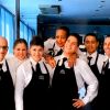Gruppo di camerieri sorridenti in uniforme con grembiule nero, posano insieme.