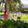 Una famiglia cammina in un parco soleggiato con palme, priva di persone intorno.