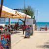 Bar sulla spiaggia con sedie sotto tende da sole, vicino al mare con bagnanti.