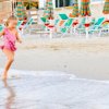 Bambina corre sorridendo verso l'acqua sulla spiaggia, mentre una donna la osserva in lontananza.