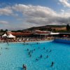 Piscina affollata con scivoli e palme, cielo parzialmente nuvoloso.