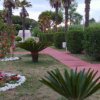 Sentiero rosso tra siepi e palme in un giardino curato con aiuole fiorite.
