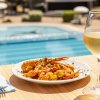 Tavolo con piatto di gamberi e pasta, bicchiere di vino bianco vicino a una piscina.