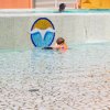 Bambino con braccioli arancioni in una piscina, accanto a un bordo con un logo a mosaico.