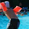 Bambino con braccioli arancioni pronto a tuffarsi in piscina affollata di nuotatori.