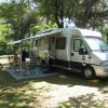 Camper parcheggiato in un'area ombrosa, con due persone sedute a un tavolo sotto un tendalino.