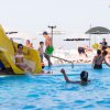 Bambini giocano in piscina con uno scivolo giallo, sotto ombrelloni bianchi e neri.