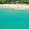 Spiaggia con ombrelloni colorati, bagnanti e mare verde chiaro. Vegetazione sullo sfondo.