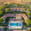 Veduta aerea di un resort con piscina e edifici circondati da alberi e campagna.
