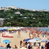 Una spiaggia affollata con ombrelloni colorati e un villaggio collinare sullo sfondo.
