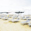 Spiaggia con sabbia chiara, sdraio grigie e ombrelloni bianchi frangiati.