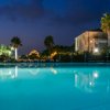 Piscina illuminata di sera con palme e edificio in pietra sullo sfondo.