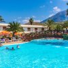Piscina con persone, palme e una passerella in legno, montagne sullo sfondo.