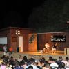 Spettacolo di cabaret serale su un palco di legno con pubblico seduto all'aperto.