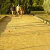 Tre persone giocano a bocce su un campo di sabbia, circondato da alberi.