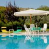 Piscina con lettini colorati e un grande ombrellone bianco, circondata da alberi e cespugli.