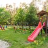 Area giochi all'aperto con scivolo rosso, altalene e alberi circostanti.