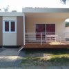 Casetta beige con terrazza e ringhiera bianca, circondata dal verde.