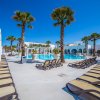 Area piscina con lettini marroni e palme, circondata da edifici bianchi sotto un cielo azzurro.