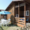 Chalet in legno con terrazza coperta, tavolo blu, sedie e ombrellone azzurro in giardino.