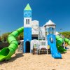 Parco giochi a tema castello con scivoli verdi e blu su sabbia, cielo limpido e alberi sullo sfondo.
