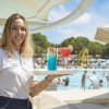 Cameriera sorridente con un vassoio di cocktail colorati davanti a una piscina affollata.